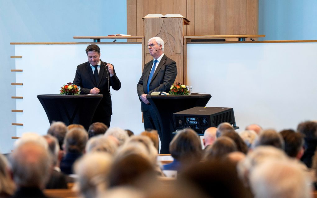 Man in zwart pak gebaart, man met wit haar luistert aandachtig en vertaalt.
