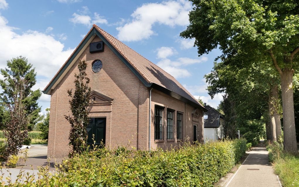 Stenen kerkgebouw met puntdak aan fietspad met bomen en heg.