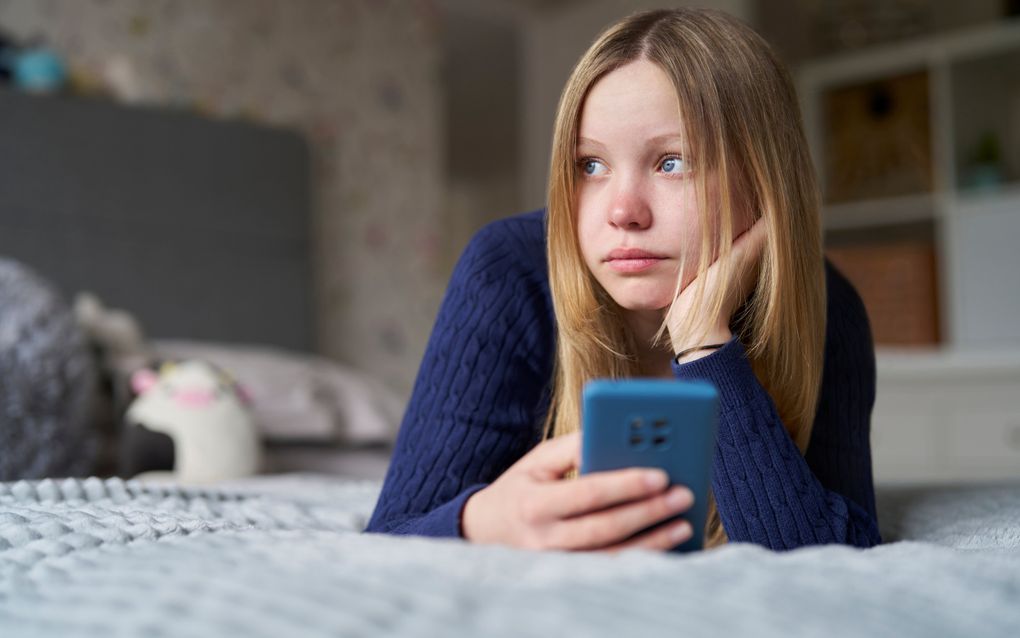 Een meisje met lang donkerblond haar en een blauwe trui ligt op haar bed met een mobiel in haar hand. Ze kijkt verdrietig.