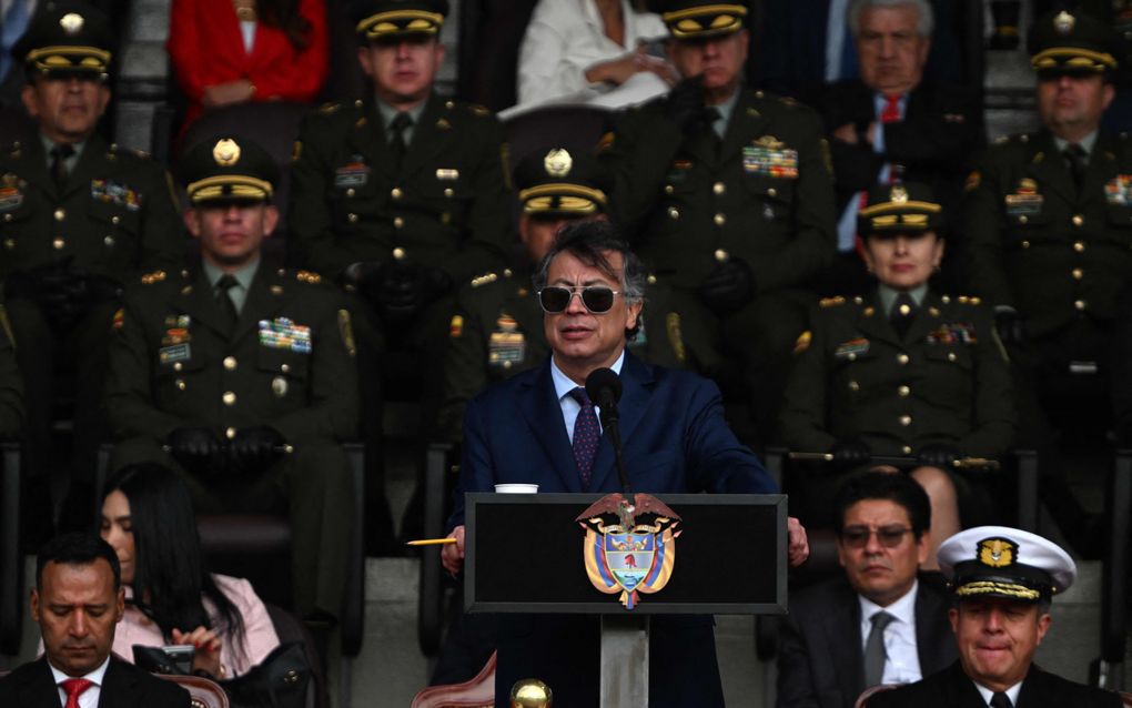 Een man in pak met een zonnebril op spreekt achter een spreekgestoelte. Achter hem staan militairen in tenue. 