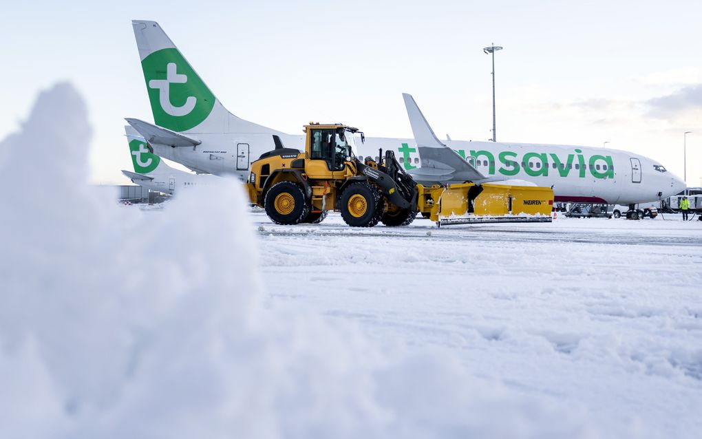 Gele sneeuwschuiver voor een witte landingsbaan waar een wit vliegtuig stilstaat.