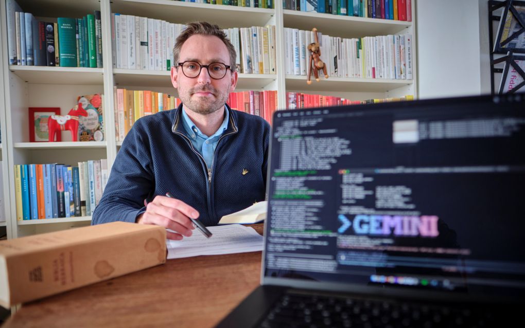 Man aan tafel met laptop en boeken om zich heen. Man draagt bril en heeft een lichte snor en baard.