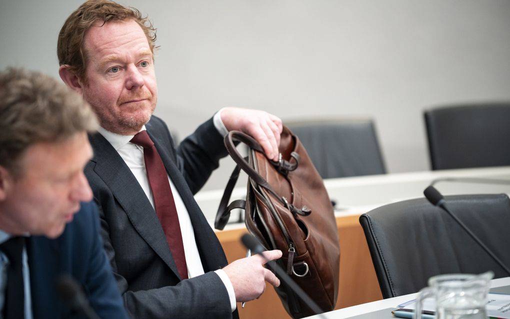 Man in donker pak, wijnrode das, met roodbruin haar en baardje, verplaatst zijn tas van de tafel op de grond. Naast hem zit een ambtenaar die de bewindsman adviseert.