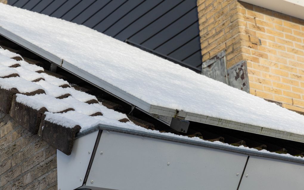 Een witte dakgoot gevuld met sneeuw, met daarboven een door sneeuw bedekt zonnepaneel.