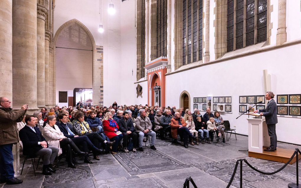 Een man in pak, achter een katheder, spreekt de aanwezigen toe in een zaal bij een kerk, met pilaren en hoge ramen.