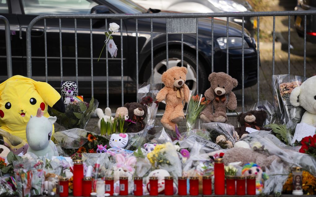Een zee aan knuffelbeertjes en bloemen op de stoep. Op de achtergrond een dranghek met erachter een zwarte auto. 