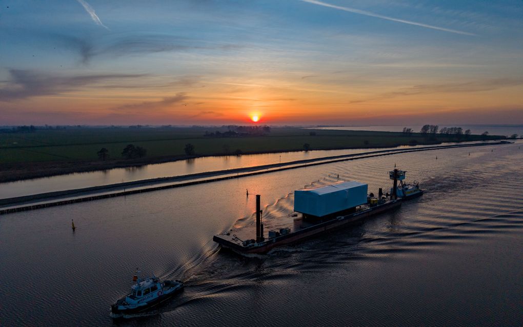 Sleepboot trekt spoor door het water. Op de achtergrond een ondergaande zon.