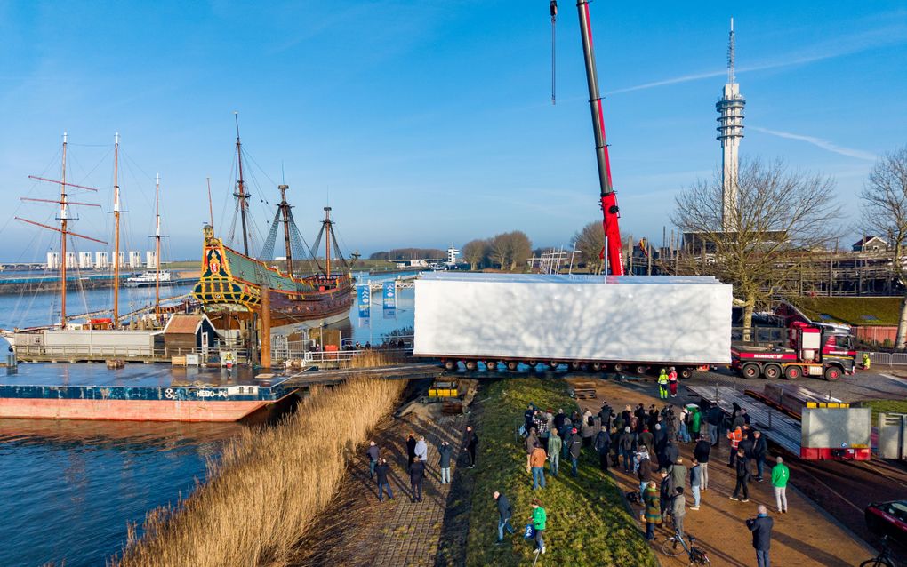 Mensen staan te kijken bij trailer die een ponton oprijdt. Links de versierde boeg van een oud schip.