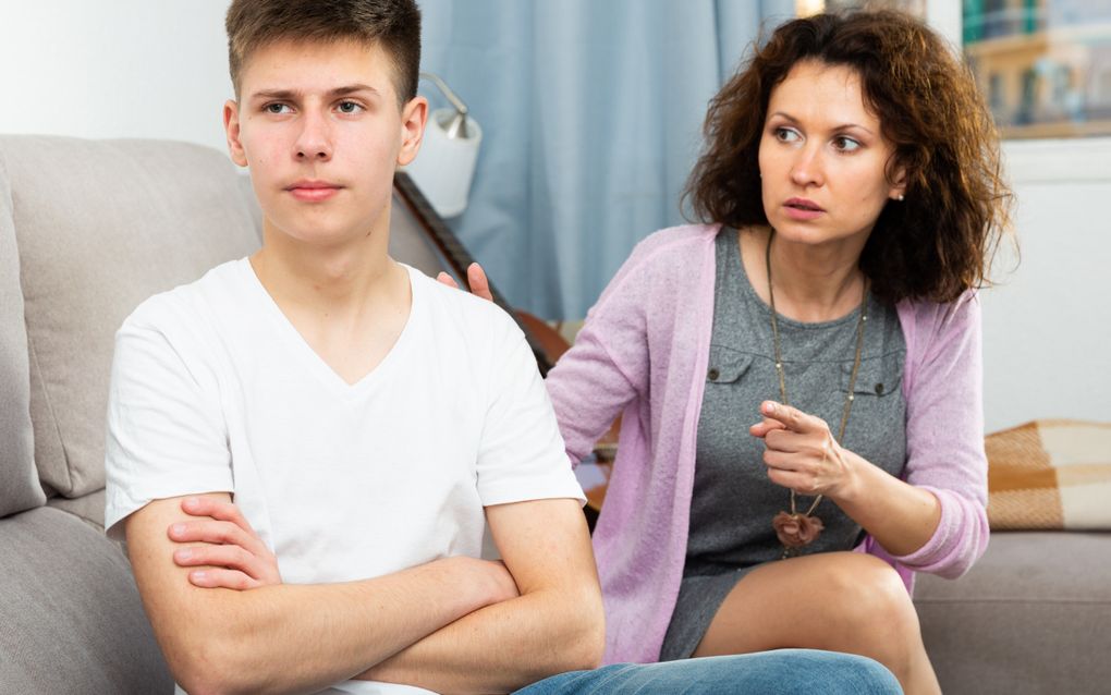 Een moeder wijst haar zoon terecht, die met zijn armen over elkaar de andere kant op kijkt.
