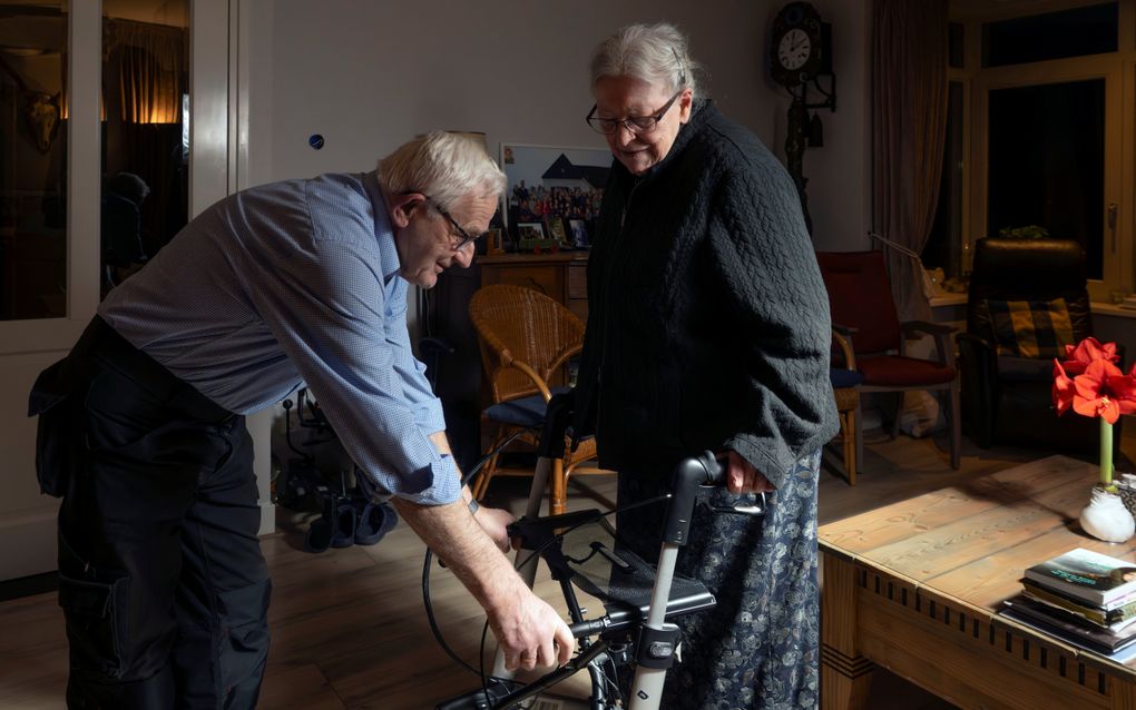 Vrouw met zwart vest staat achter rollator, man met blauw overhemd buigt voorover om de rollator vast te houden.