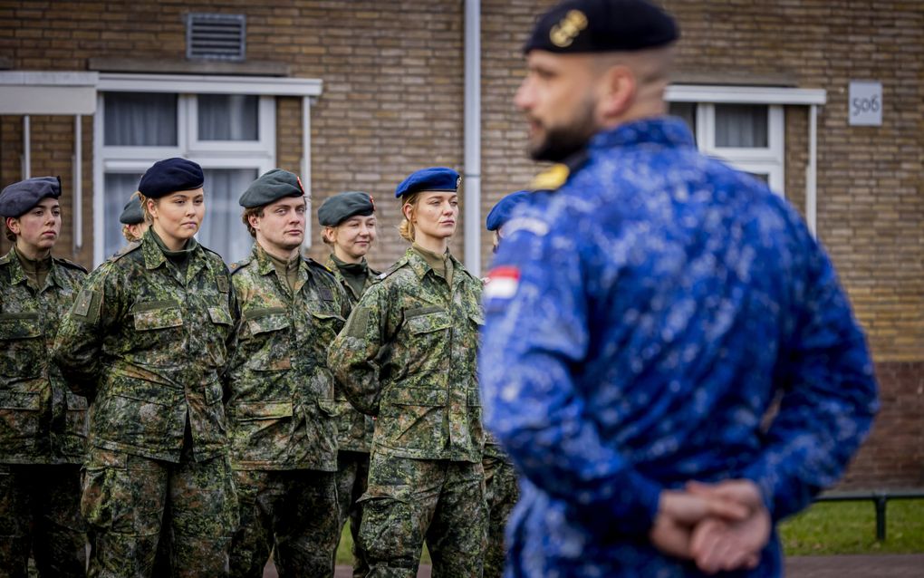 Man in blauw marine-uniform staat met de handen op de rug voor een groep jonge militairen-in-opleiding. Zij dragen een groen uniform en een baret.