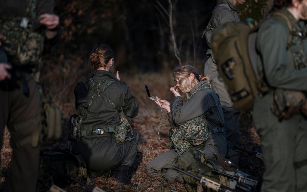 Jonge vrouw in militair uniform zit op haar knieën in het bos, terwijl ze met één hand donkergroene schmink op haar gezicht smeert. In de andere hand houdt ze een spiegeltje.