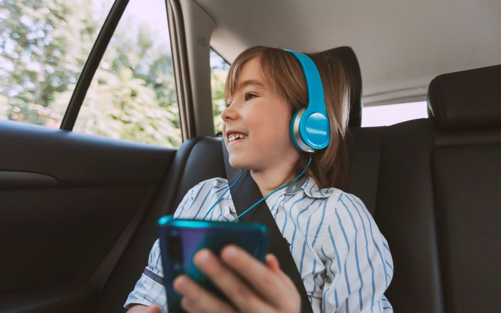 Een meisje zit op de achterbank van een auto, in de gordel, met een koptelefoon op een een smartphone in haar hand.