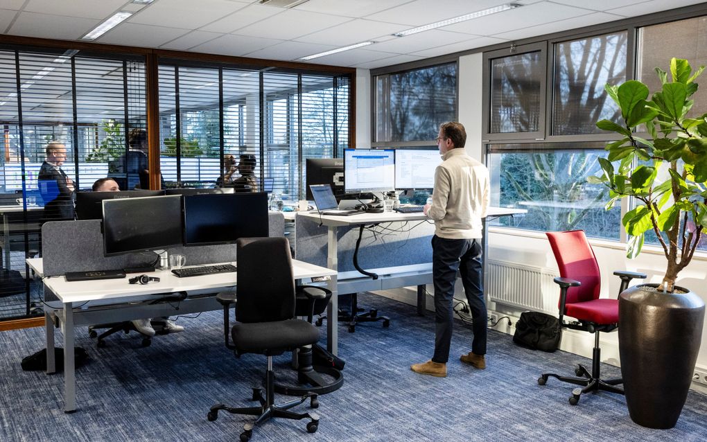 Een kantooromgeving met diverse bureaus, schermen en stoelen. Rechts taat een man achter een hoog bureau, achter hem een rode stoel. Bij een ander bureau staat een ergonomische stoel. Dat is meer een zitvlak met een verende poot eronder.