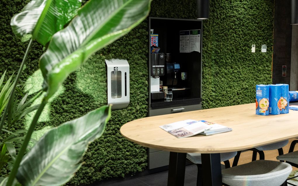Kantoorruimte met links een groene plant, rechts een tafel met daarop blauwe verpakking met frisdrank en wat tijdschriften. Op de achtergrond een is in de wand met daarin een drankenautomaat.