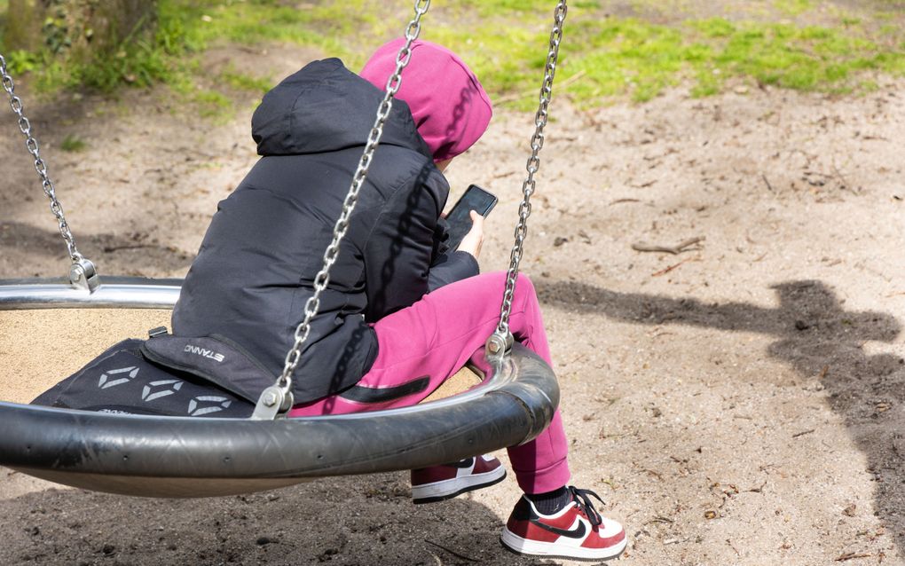 Jongere met zwart jack en roze broek op schommel, met mobiele telefoon in de hand