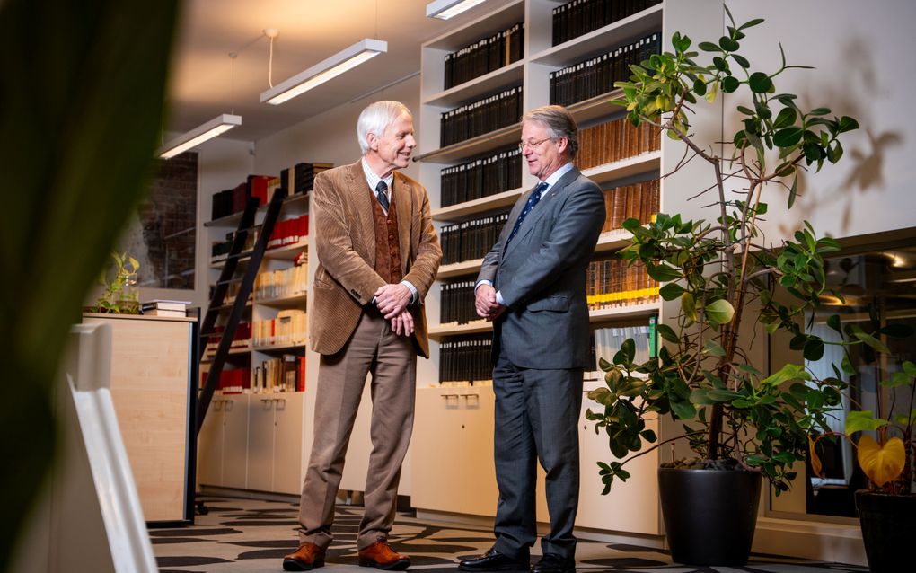 Twee grijzende mannen, gestoken in een pak, staan te praten. Achter hen staat een kast vol met boeken. Ter rechterzijde van de mannen staat een plant met groene bladeren.
