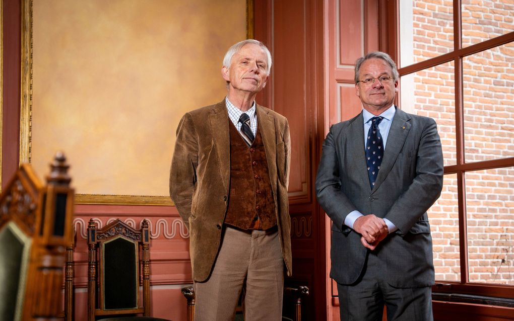 Twee grijsharige mannen in pak staan in een kamer met rode lambrisering en twee ouderwetse stoelen van hout.