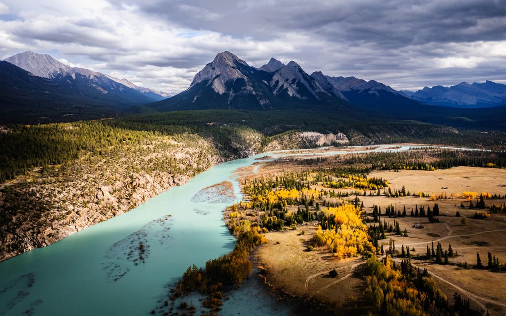 Groenige rivier stroomt door een met zonlicht beschenen landschap. Aan de horizon zijn besneeuwde bergtoppen te zien.