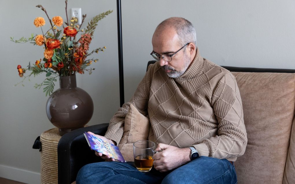 Kalende man in bruine coltrui en spijkerbroek zit in een bruine stoel met een boekje op schoot en een kop thee in zijn hand. Rechts van hem stat een bruine vaas met oranje bloemen erin.