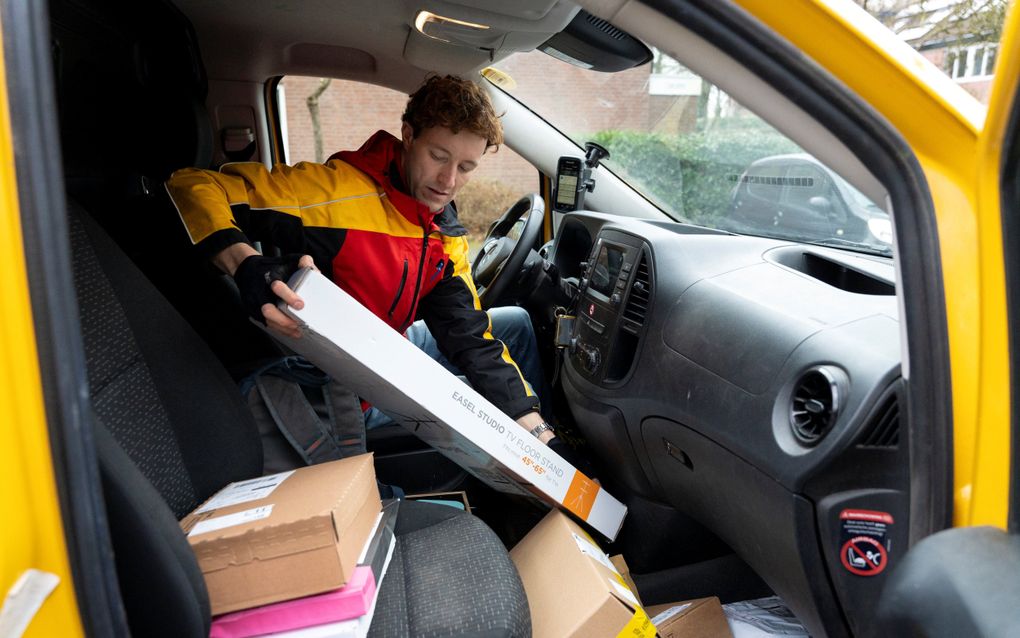 Een jongeman met geelrode jas. Hij zit in een auto achter het stuur en houdt een pakketje vast. 