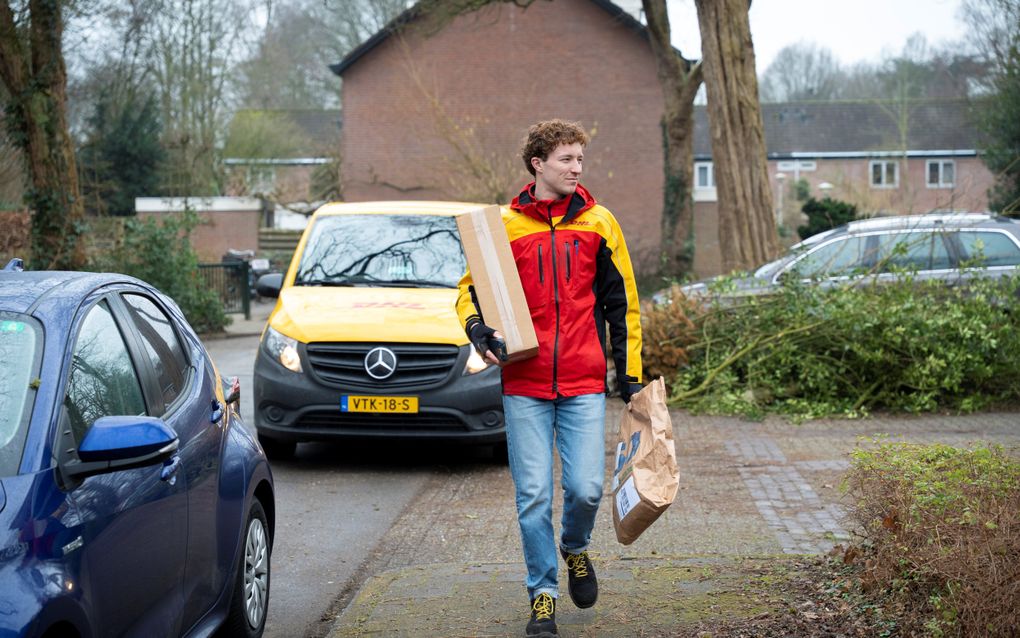 Jongeman in geelrode jas, wandelt met twee pakketjes in de hand over een stoep. 