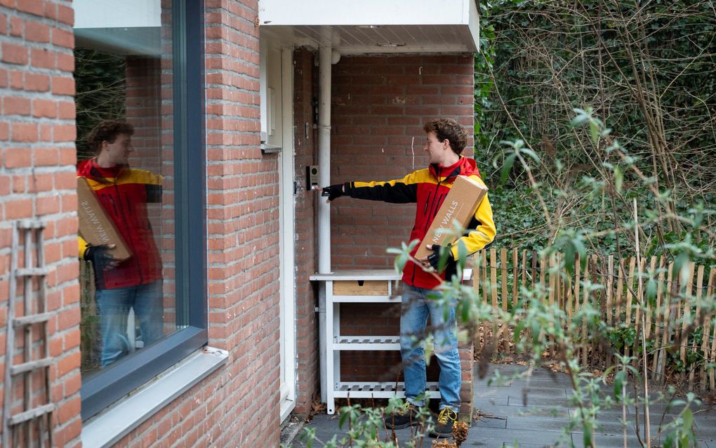 Een man met geelrode jas en een pakket in zijn arm belt aan bij een huis. 