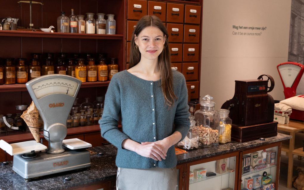 Jonge vrouw staat voor een toonbank, waarop een ouderwetse weegschaal en kassa staan. Achter haar tegen de muur staat een grote medicijnkast.