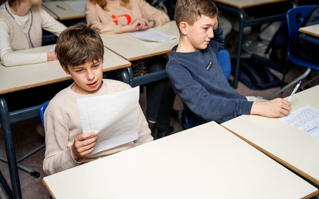 Een jongen met donker haar leest iets voor van een wit vel papier. Naast hem zit een andere jongen.