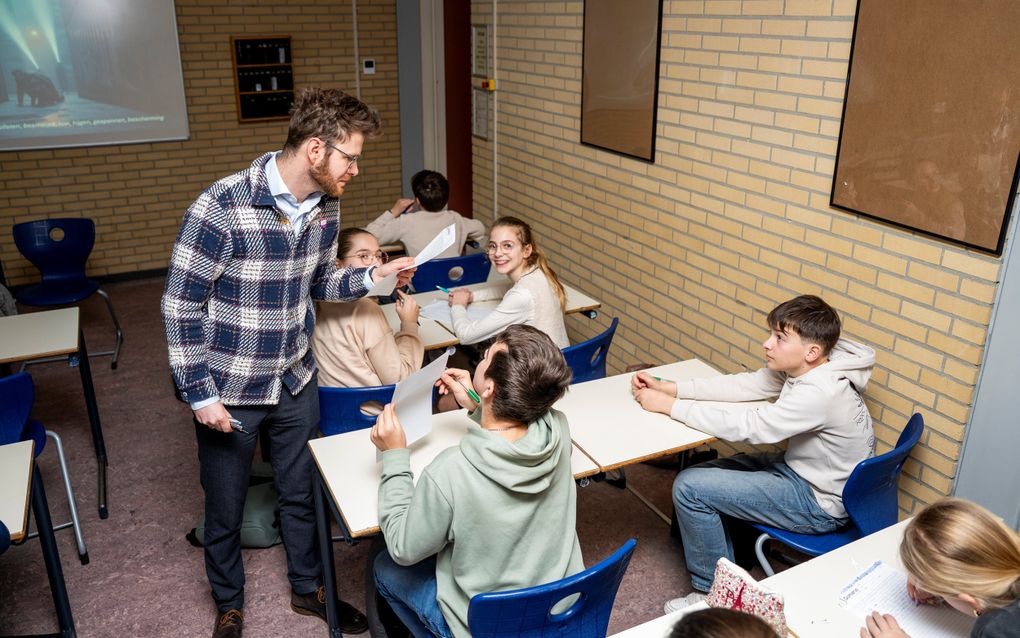 Een man kijkt naar jongens en meisjes die op stoelen achter tafels zitten.