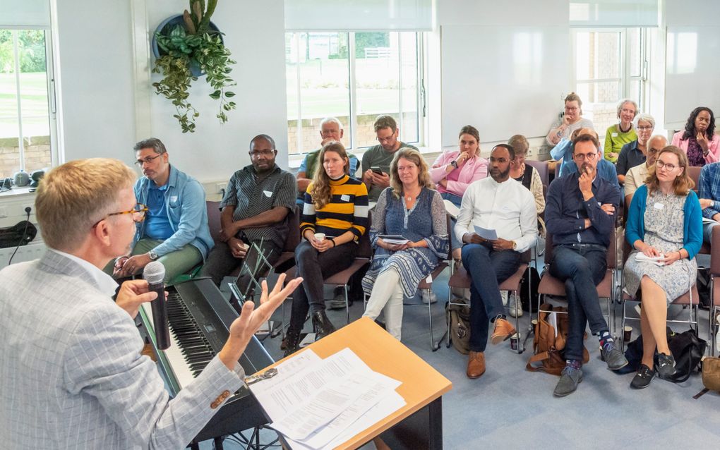 Man achter een katheder spreekt een zaaltje met mensen toe.
