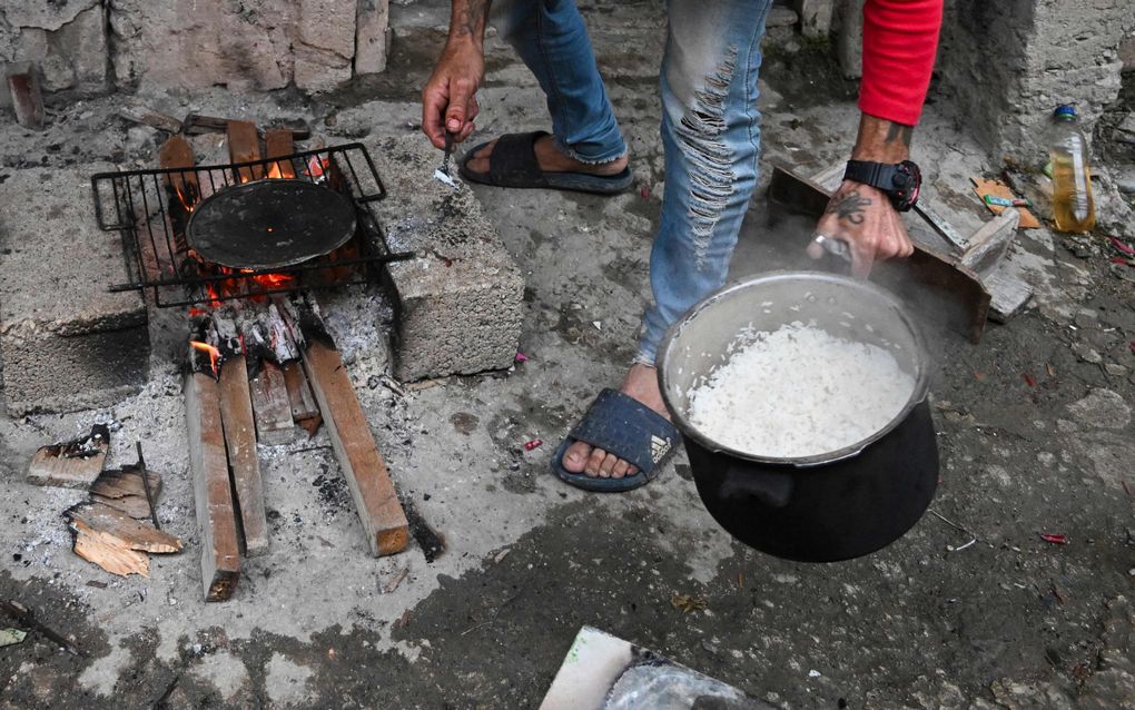 Een hand houdt een pan rijst vast. Naast de voeten van een man brandt een vuurtje. 