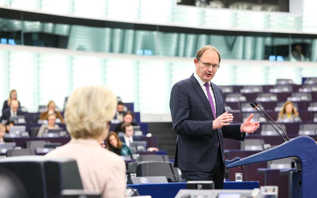 Bebrilde man in donkerblauw pak en met paarse das spreekt richting een blonde vrouw. Hij gebaart met zijn handen.