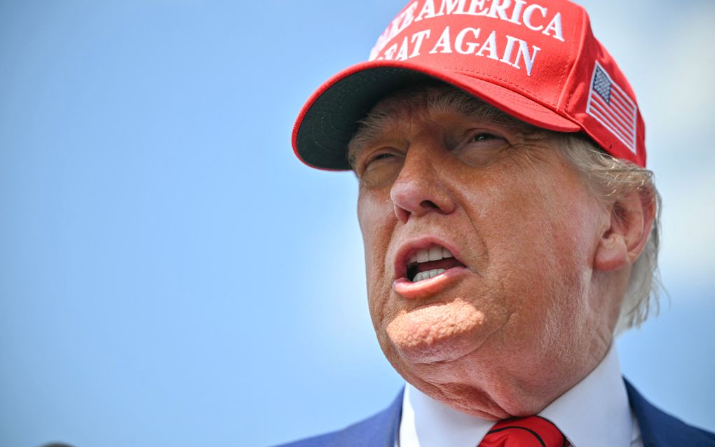Foto van president Trump met een rode MAGA-pet op, tegen een blauwe lucht.