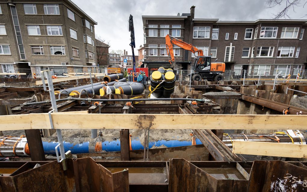 Voor een rij woonhuizen ligt een open bouwput, waarvan de wanden gestut zijn met damwand. Op stalen balken liggen kabels en buizen.