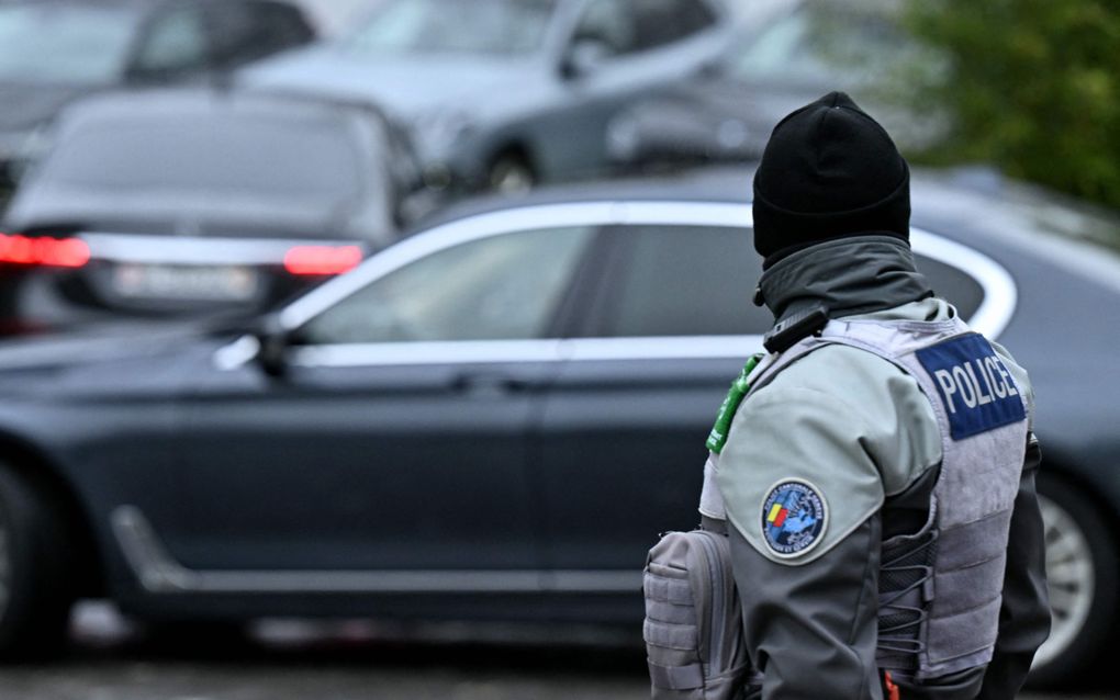 Politieagent in grijs uiform met muts op staat op straat, terwijl donkergekleurde auto’s passeren.