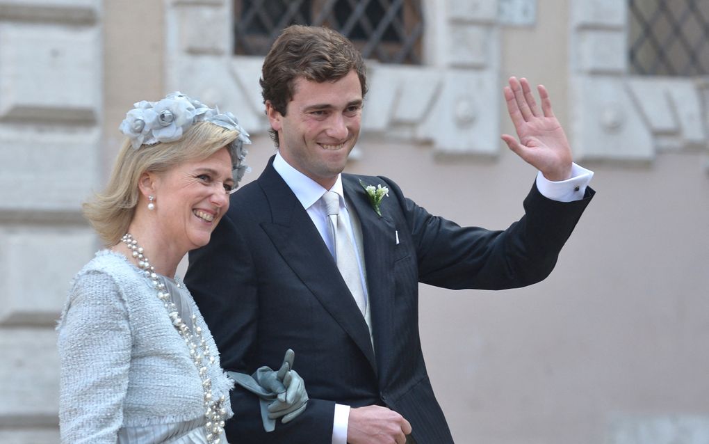 Jonge man met golvend bruin haar, in donker pak met wit overhemd, corsage op de revers, zwaait. Aan zijn arm zijn moeder, met handschoenen in haar hand.