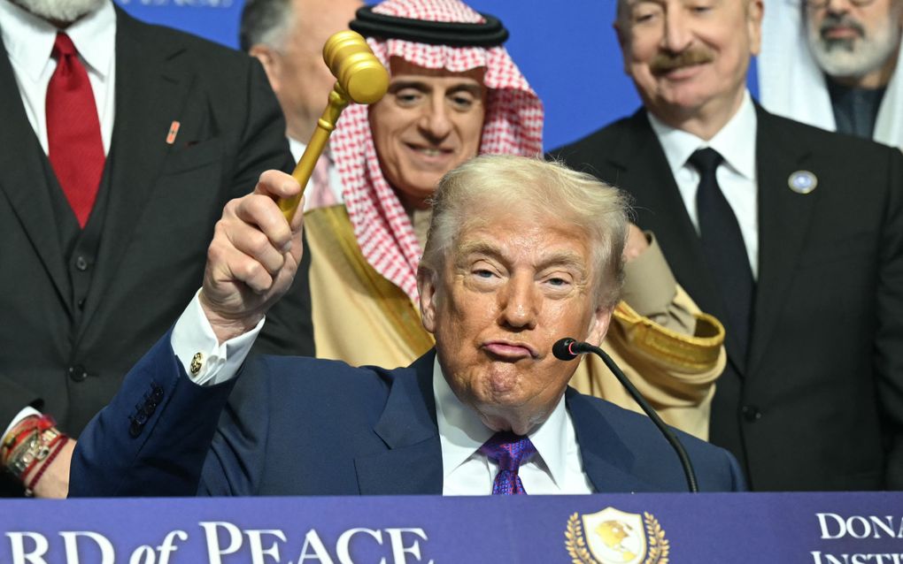 Een blonde man in pak houdt een voorzittershamer in de lucht. Achter hem staat een Arabische man.