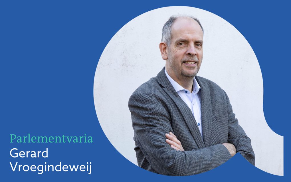 Foto van een glimlachende en wat oudere, kalende man in blauwgrijs colbert, zonder stropdas en met armen over elkaar.