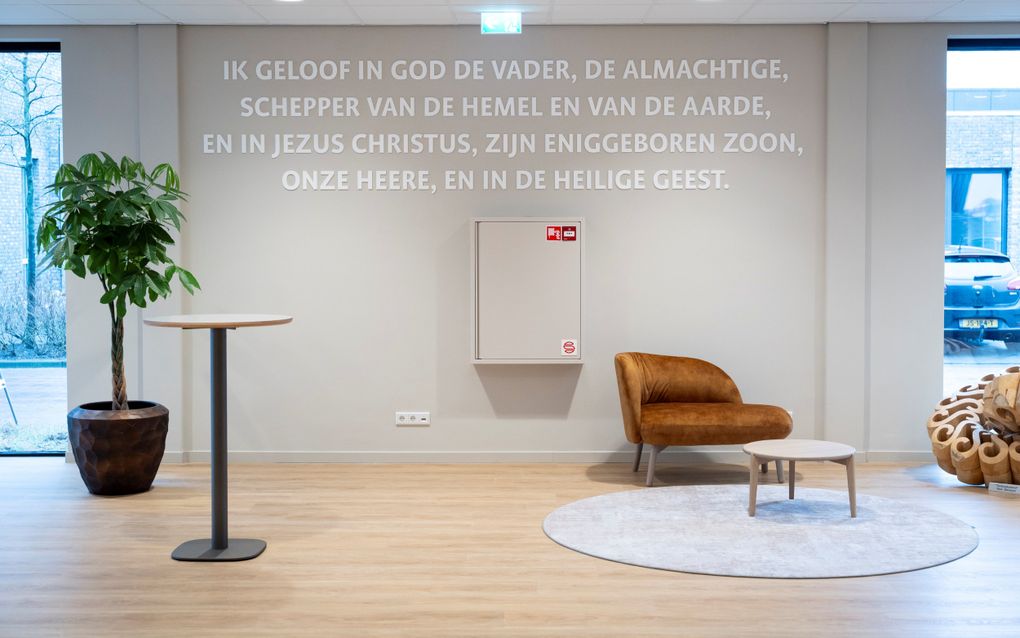 Tekst op de muur in de hal: „Ik geloof in God de Vader…”. Ervoor een modern vormgegeven stoel.