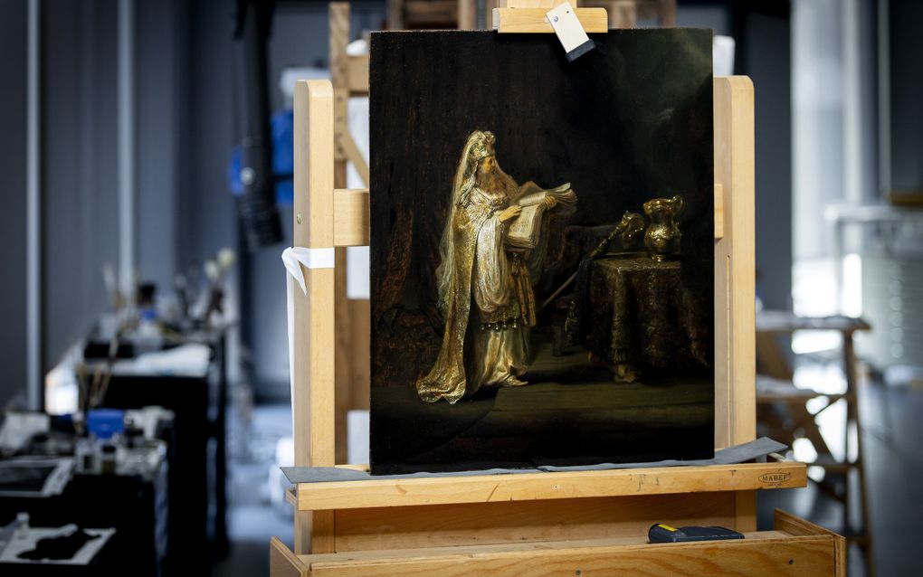 Foto van een schilderij van een oude man in goudkleurig oosters gewaad en met een opengeslagen boek in zijn hand. Het schilderij staat op een ezel in een atelier.