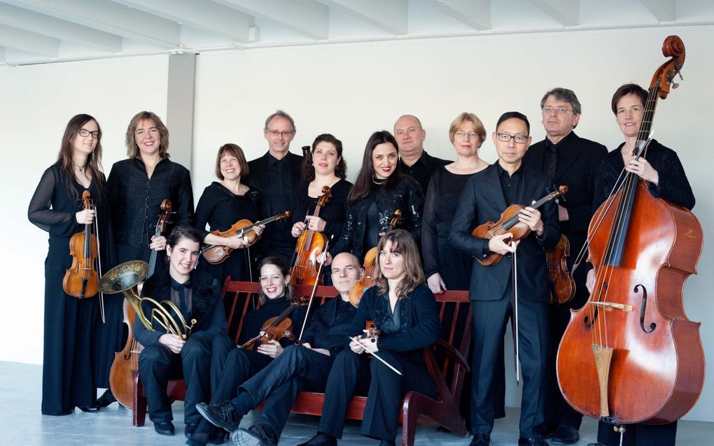 Groep mensen in zwarte kleren met allerlei muziekinstrumenten voor zich. Een deel zit op een bank, een ander deel staat achter en om de bank heen. 
