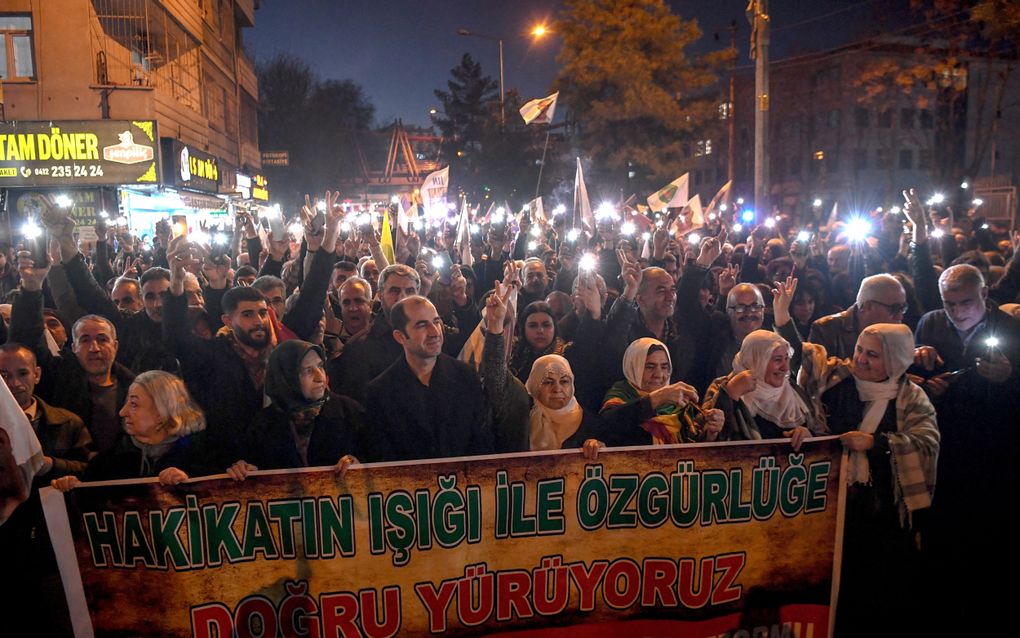 In het donker staan honderden mensen op straat te zwaaien met zaklampjes en vlaggen. Vrouwen met hoofddoek lopen vooraan met een groot protestbord in hun handen.