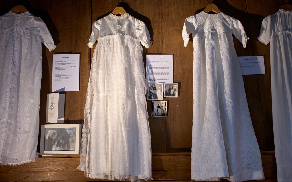 Een viertal doopjurken, voorzien van een toelichting, aan een houten wand. Er hangen ook enkele oude foto’s.