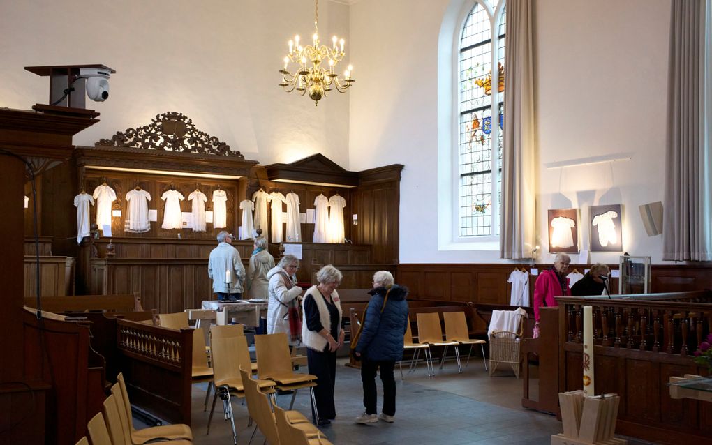 Oudere bezoekers lopen door de kerkruimte waarin de doopjurken te zien zijn.