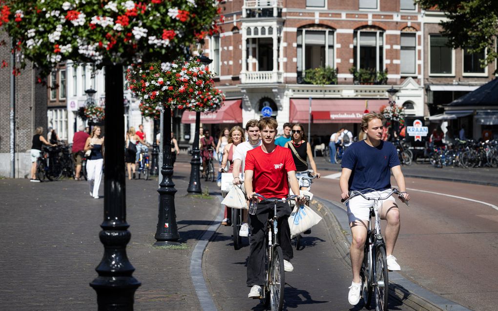 Een aantal jonge mensen in zomerse kleding op de fiets. Naast de weg staan lantaarnpalen met bloembakken; de bloemen zijn in de kleuren rood en wit.