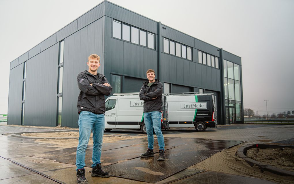 Twee jongemannen die allebei een jack en een spijkerbroek dragen, staan voor een grijs bedrijfspand en een wit busje.