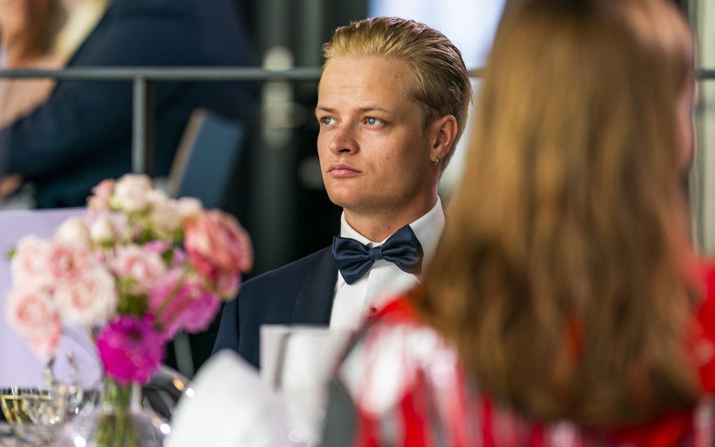 Een blonde jongen in jacquet zit aan een tafel waarop bloemen staan.