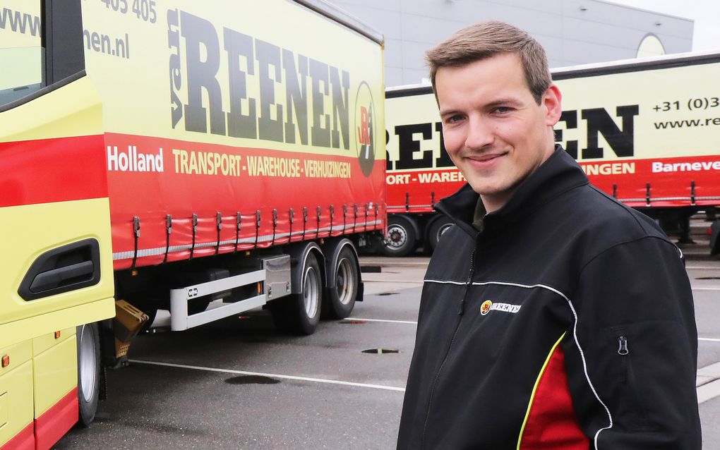 Een jongeman met zwart-rode jas staat voor twee rood-gele vrachtwagens van Van Reenen Transport.