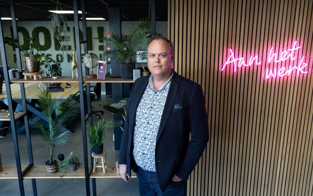 Forse man in een bewerkt overhemd en donker colbert staat voor een wand waarop in roze letters „aan het werk” staat.